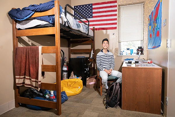 A young man sits at his desk in a narrow dorm room, his bunk bed behind him and an American flag on the wall above.