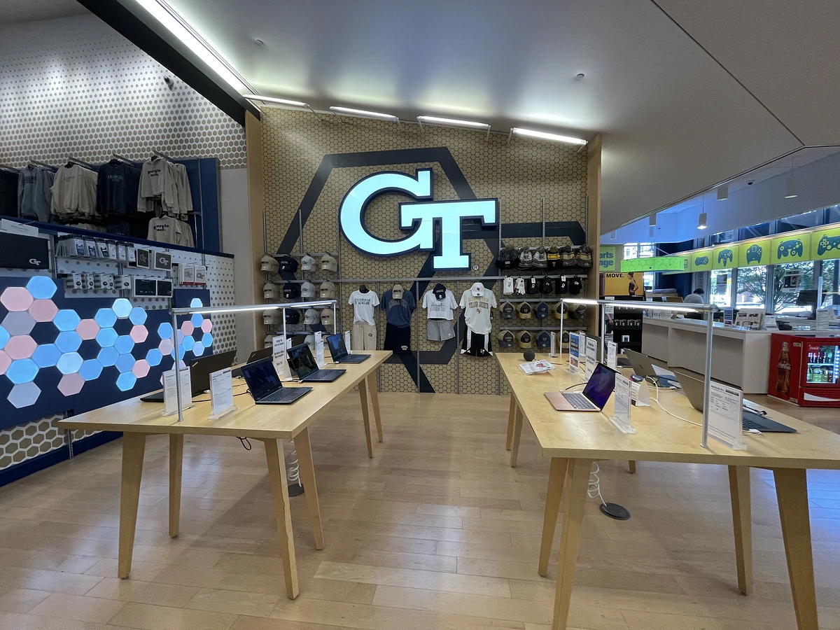 A tech-oriented retail space features laptops on display and branded merchandise against a backdrop with a prominent "GT" logo.
