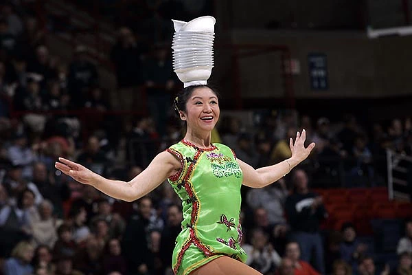 A performer in a vibrant green outfit balances a stack of bowls on her head while entertaining a crowd.