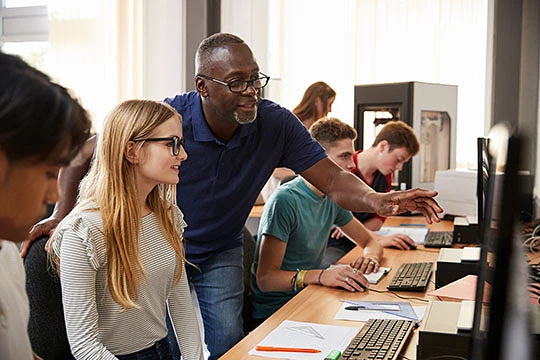 A teacher is assisting students working on computers in a classroom setting.